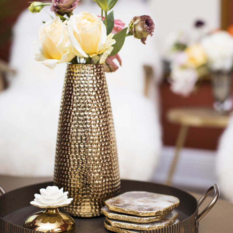 closeup photo of hammered gold vase with flowers, gold coasters on antique brass tray