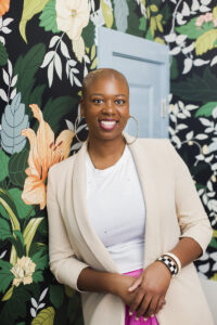 Whitney Jones, founder and principal designer of Whitney J Decor, smiling in a bright studio setting. She is wearing a stylish outfit and standing confidently, reflecting her expressive, color‑driven design style.