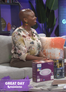 Interior designer Whitney Jones on Great Day Louisiana discussing lighting and home décor products, seated on a studio couch with featured Philips and Hampton Bay lighting items displayed on the table.